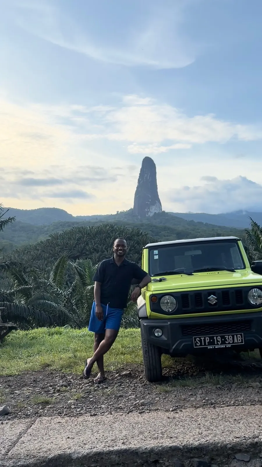 Me leaning on a lime-green Suzuki Jimny with the Pico Cão Grande peak rising behind me in São Tomé